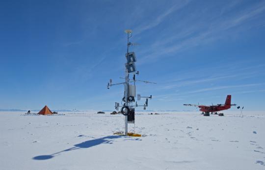 Photo of a tall scientific instrument sticking out the snow, with distant pyramid tent to the left, and distant twin otter aircraft to the right.