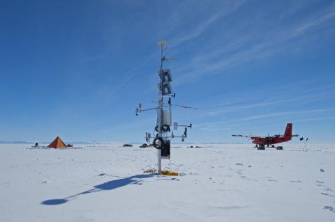 Photo of a tall scientific instrument sticking out the snow, with distant pyramid tent to the left, and distant twin otter aircraft to the right.