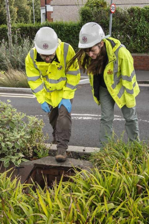 Two people in hard hats and hi-vis jackets peer into a hole in the ground.